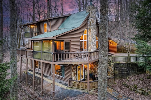 back of house at dusk with log veneer siding, metal roof, a chimney, and a wooden deck