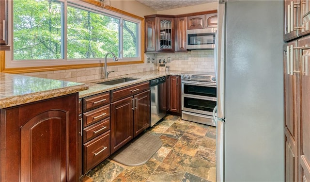 kitchen with tasteful backsplash, stone finish floor, appliances with stainless steel finishes, glass insert cabinets, and a sink