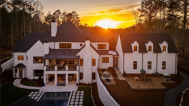 back of house at dusk featuring a balcony, a chimney, metal roof, a standing seam roof, and a patio area