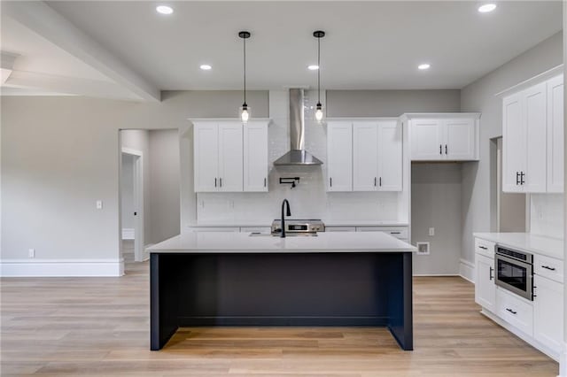 kitchen with decorative backsplash, light countertops, light wood-style flooring, a kitchen island with sink, and wall chimney exhaust hood