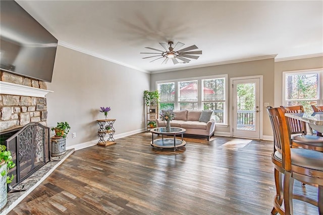 living area featuring a stone fireplace, wood finished floors, a ceiling fan, baseboards, and crown molding