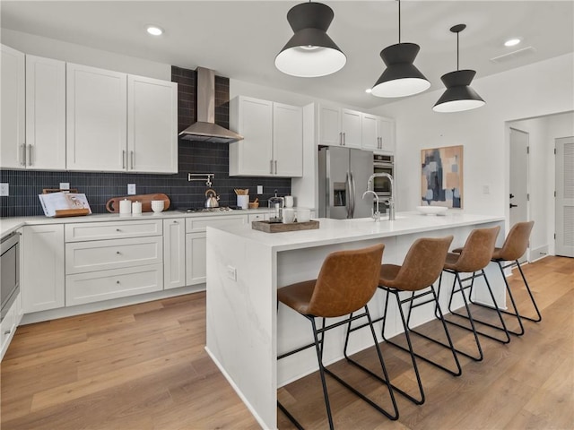 kitchen featuring stainless steel appliances, light wood-type flooring, wall chimney range hood, and tasteful backsplash