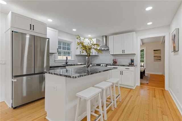 kitchen featuring a center island, wall chimney range hood, dark stone countertops, white cabinets, and stainless steel fridge