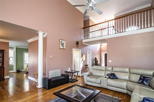 living room featuring a high ceiling, wood-type flooring, decorative columns, ornamental molding, and ceiling fan