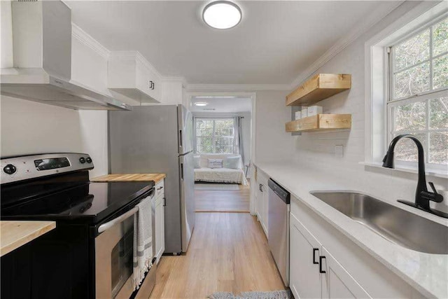 kitchen with wall chimney range hood, appliances with stainless steel finishes, a sink, white cabinets, and light wood finished floors