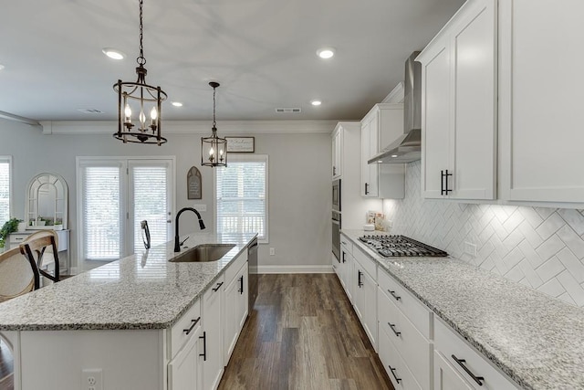 kitchen featuring sink, hanging light fixtures, an island with sink, wall chimney range hood, and white cabinets