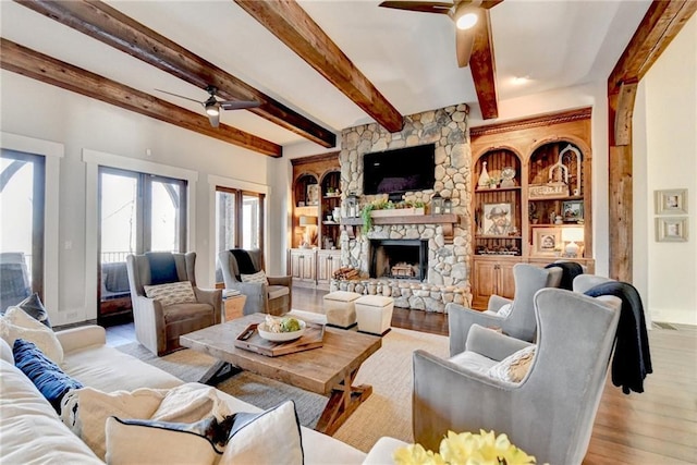 living room featuring beamed ceiling, built in shelves, a ceiling fan, a stone fireplace, and light wood finished floors