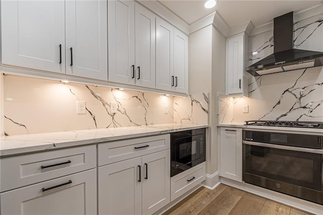 kitchen with wall chimney exhaust hood, white cabinetry, light stone countertops, light wood-type flooring, and black appliances