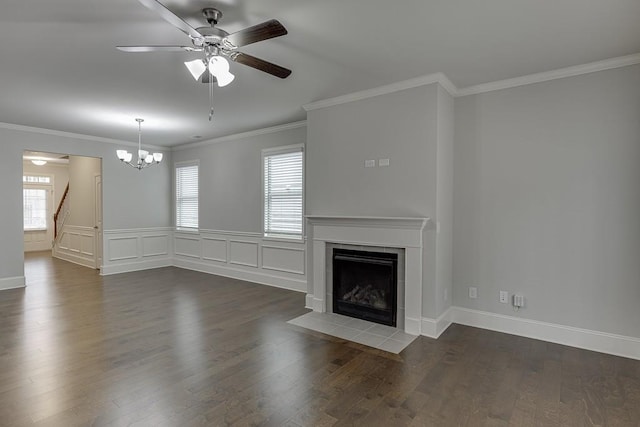 unfurnished living room with ceiling fan with notable chandelier, dark wood-style flooring, wainscoting, a tiled fireplace, and crown molding