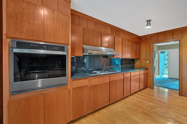 kitchen featuring under cabinet range hood, stainless steel appliances, backsplash, light wood-style flooring, and dark countertops
