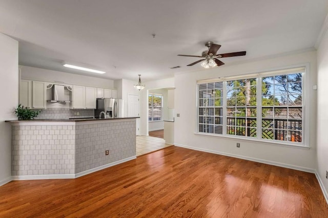 kitchen with wall chimney range hood, stainless steel refrigerator with ice dispenser, decorative light fixtures, light wood-type flooring, and white cabinetry