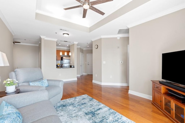 living area featuring a ceiling fan, light wood-style floors, a tray ceiling, ornamental molding, and baseboards