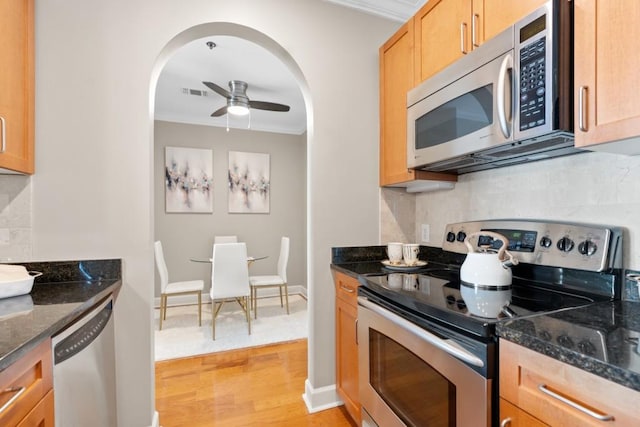 kitchen with stainless steel appliances, ornamental molding, dark stone counters, backsplash, and light wood-style floors
