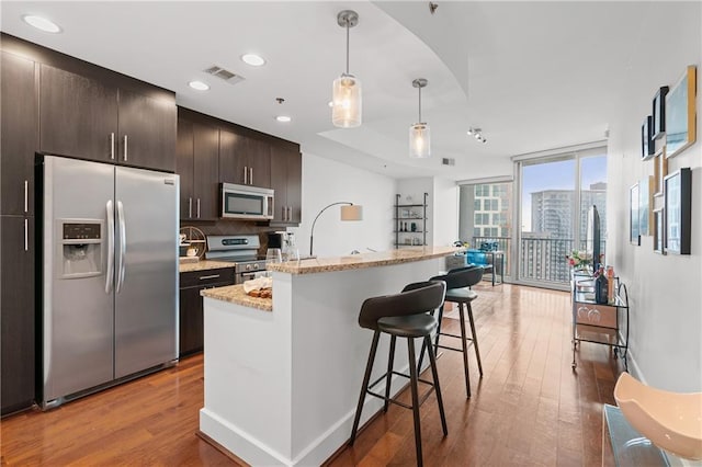 kitchen featuring stainless steel appliances, floor to ceiling windows, dark brown cabinetry, wood-type flooring, and an island with sink