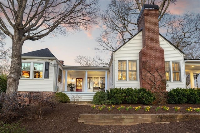 back of house at dusk featuring covered porch and a chimney