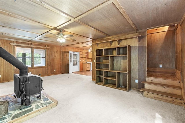 unfurnished living room featuring wooden ceiling, stairway, wood walls, a wood stove, and a ceiling fan