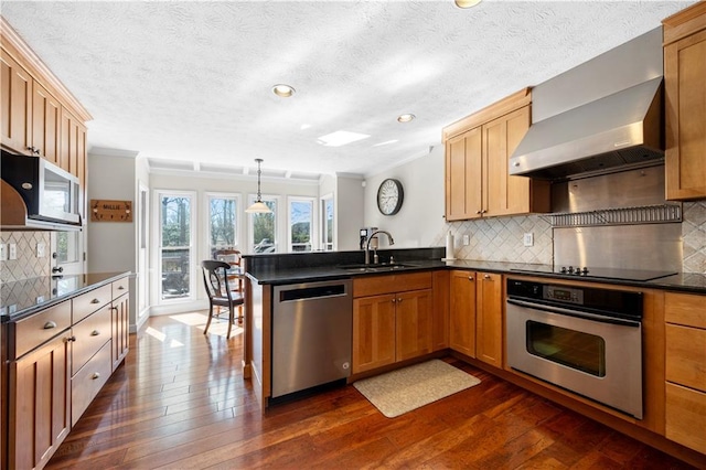 kitchen featuring stainless steel appliances, a peninsula, a sink, wall chimney range hood, and dark countertops