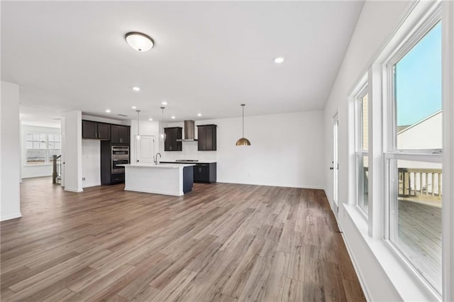 kitchen featuring a healthy amount of sunlight, wall chimney exhaust hood, hanging light fixtures, and a center island with sink