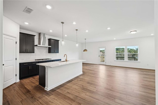 kitchen with pendant lighting, stainless steel gas stovetop, an island with sink, sink, and wall chimney exhaust hood