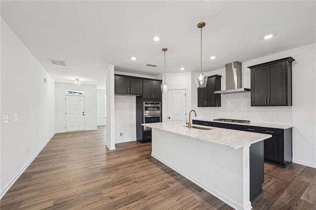 kitchen featuring sink, stainless steel appliances, light stone countertops, a center island with sink, and wall chimney exhaust hood