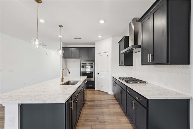 kitchen with an island with sink, sink, hanging light fixtures, stainless steel appliances, and wall chimney exhaust hood