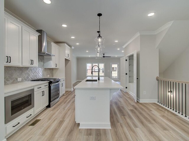 kitchen featuring white cabinets, wall chimney range hood, stainless steel appliances, hanging light fixtures, and a center island with sink