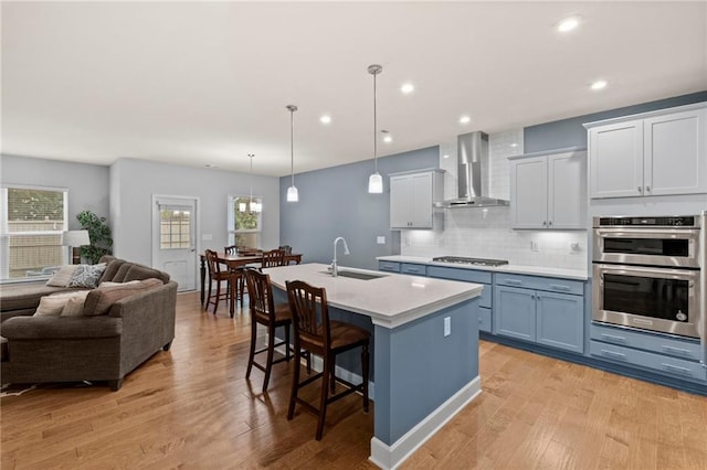 kitchen with wall chimney range hood, a sink, open floor plan, light countertops, and an island with sink
