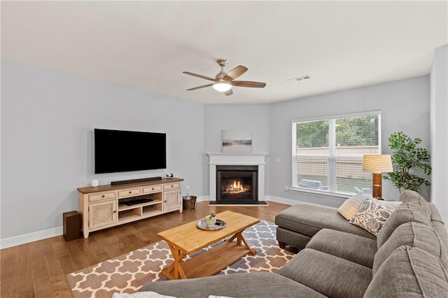 living room featuring a ceiling fan, dark wood finished floors, a warm lit fireplace, and baseboards