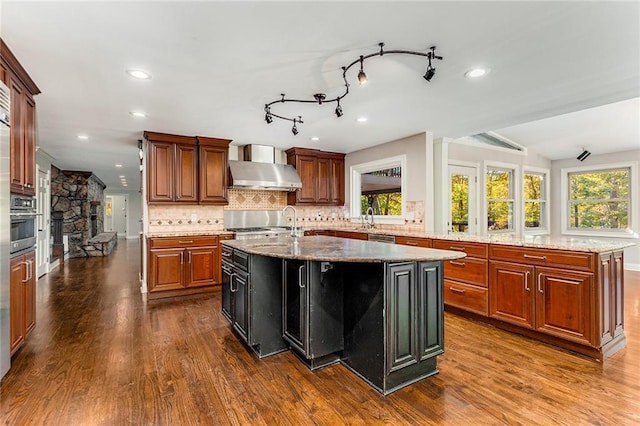 kitchen featuring ventilation hood, an island with sink, light stone countertops, and backsplash