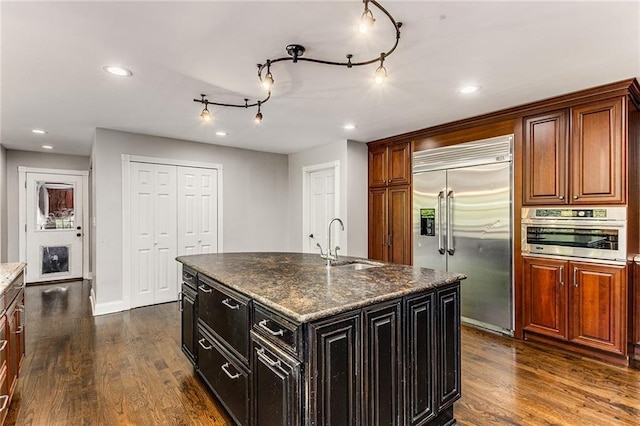 kitchen featuring sink, appliances with stainless steel finishes, a center island with sink, dark hardwood / wood-style flooring, and dark stone counters