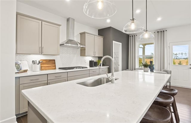 kitchen with pendant lighting, wall chimney range hood, sink, and a center island with sink