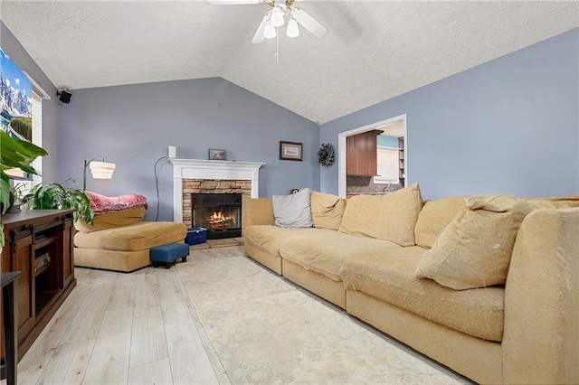 living room featuring light wood-style flooring, ceiling fan, vaulted ceiling, a textured ceiling, and a stone fireplace