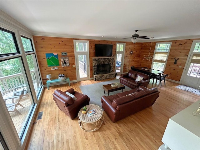 living room featuring wood walls, a stone fireplace, ceiling fan, ornamental molding, and light hardwood / wood-style flooring