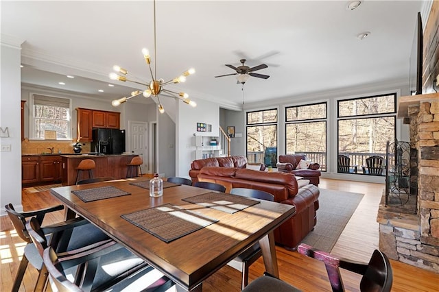 dining area with light wood-type flooring, crown molding, a stone fireplace, and ceiling fan with notable chandelier