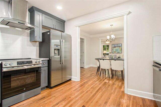 kitchen with wall chimney range hood, stainless steel appliances, a chandelier, baseboards, and light wood-style floors