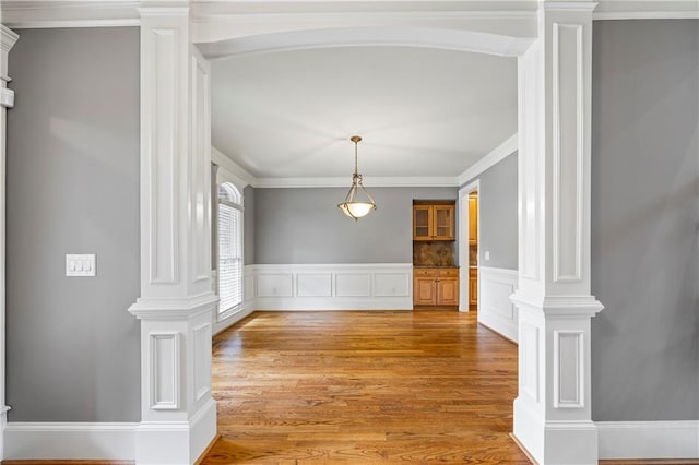 unfurnished dining area with decorative columns, crown molding, a decorative wall, light wood-type flooring, and wainscoting