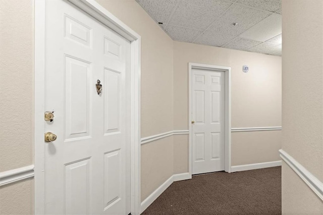 corridor featuring a paneled ceiling, dark carpet, and baseboards