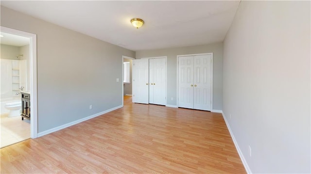 unfurnished bedroom featuring ensuite bath, light wood-style flooring, two closets, and baseboards