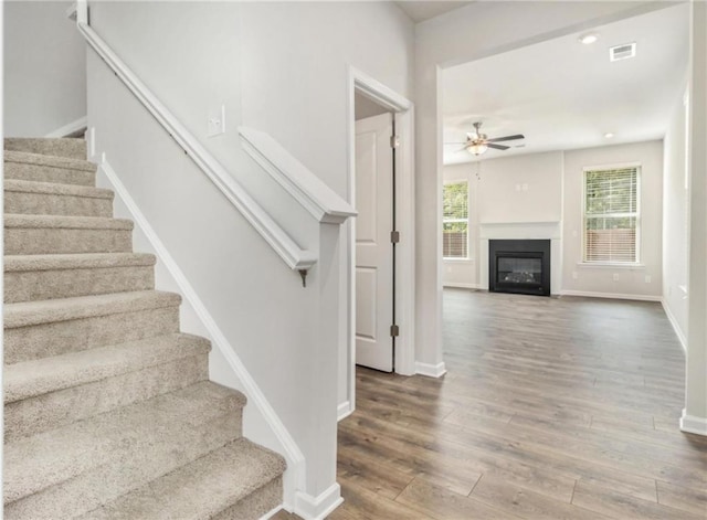 stairway with ceiling fan, a glass covered fireplace, visible vents, wood finished floors, and baseboards