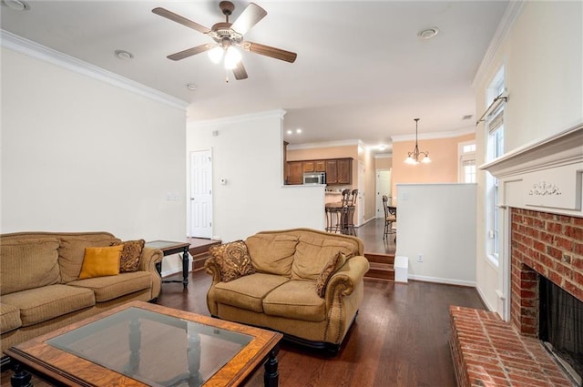 living area with baseboards, dark wood-type flooring, crown molding, a brick fireplace, and ceiling fan with notable chandelier