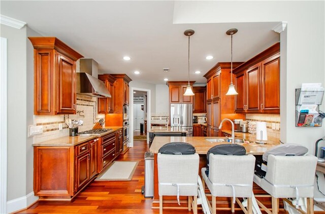 kitchen with appliances with stainless steel finishes, a sink, light wood finished floors, a peninsula, and wall chimney exhaust hood