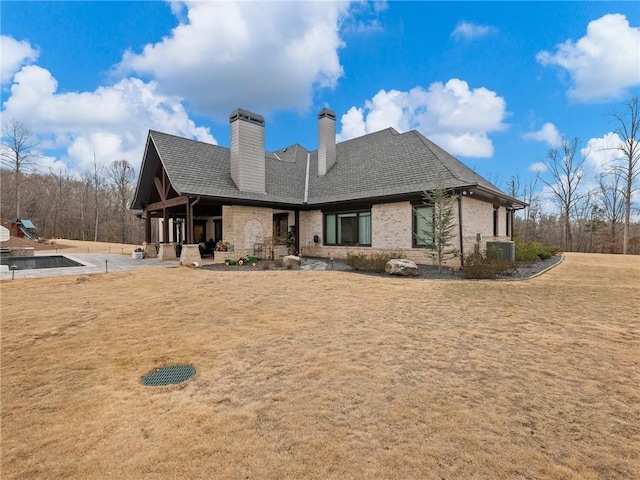 rear view of property with cooling unit, roof with shingles, a chimney, a lawn, and brick siding