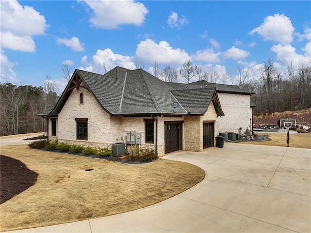 french provincial home with driveway, cooling unit, an attached garage, a shingled roof, and brick siding