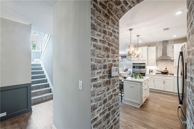 kitchen with stainless steel double oven, wall chimney exhaust hood, a kitchen island, decorative light fixtures, and white cabinetry