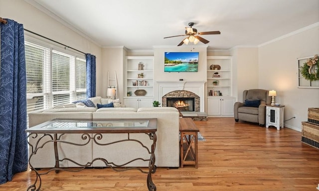 living area with light wood finished floors, crown molding, built in features, a warm lit fireplace, and a ceiling fan