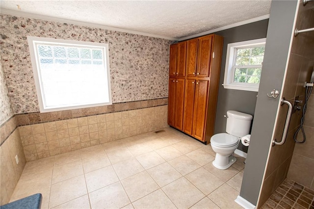 bathroom featuring a textured ceiling, toilet, ornamental molding, tiled shower, and a wainscoted wall