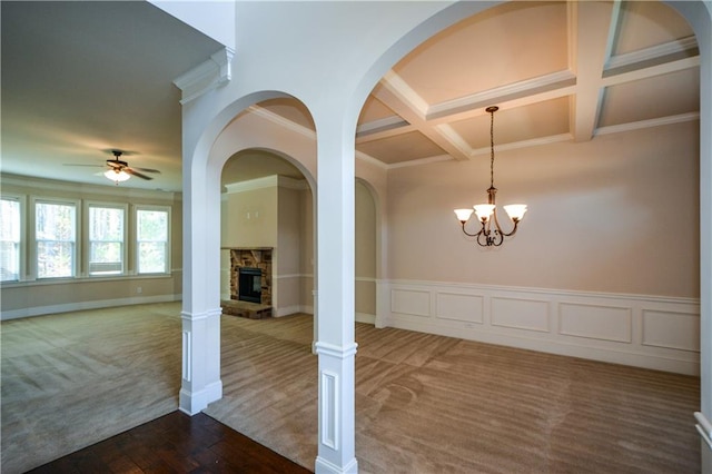 interior space with carpet, coffered ceiling, ceiling fan with notable chandelier, a stone fireplace, and crown molding