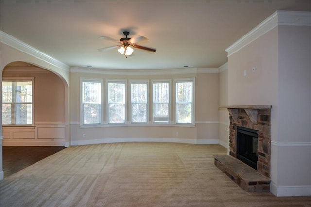 unfurnished living room with ceiling fan, a stone fireplace, light colored carpet, and crown molding