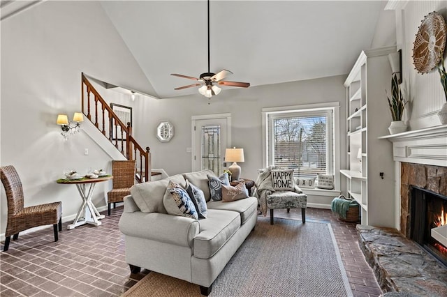 living area featuring brick floor, a ceiling fan, baseboards, stairs, and a lit fireplace