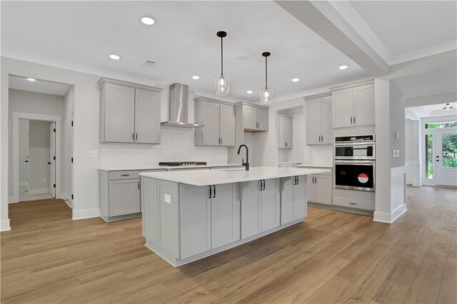 kitchen featuring double oven, wall chimney range hood, an island with sink, and light hardwood / wood-style flooring